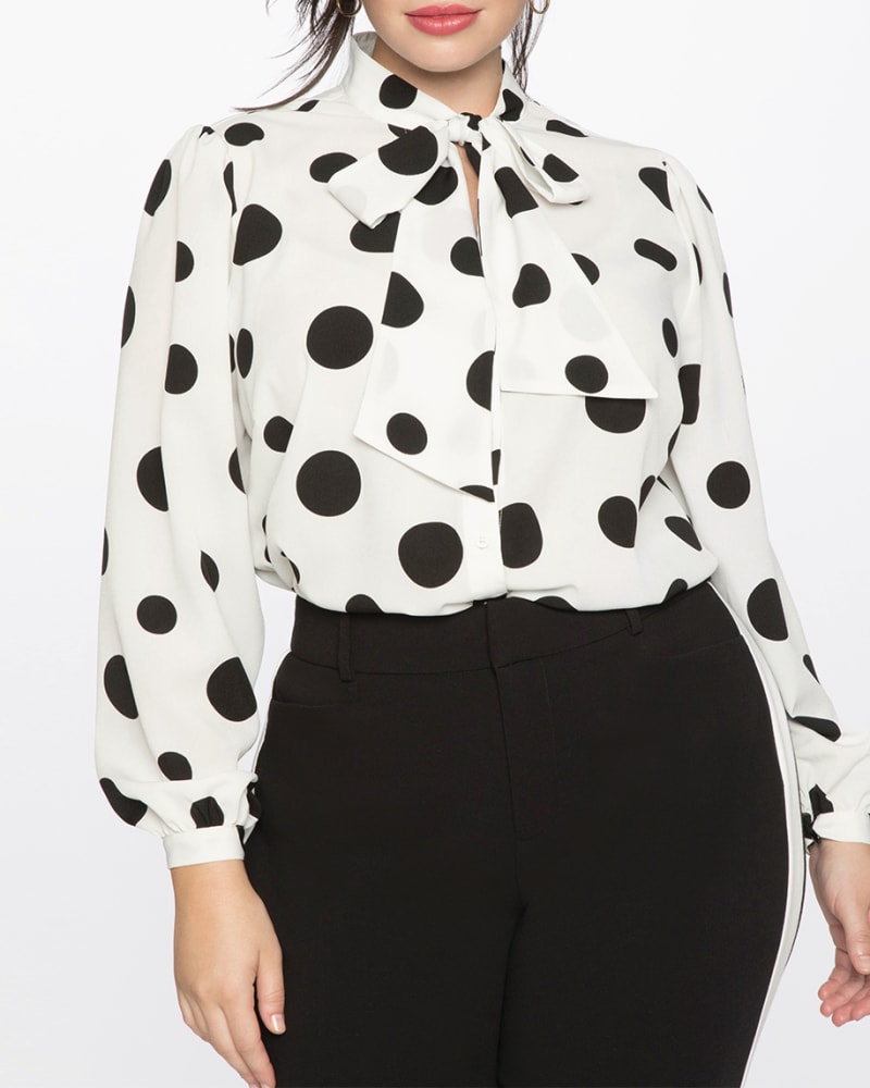 Front of a model wearing a size 14 Reina Tie Neck Blouse in Soft White Ground With Black Polka Dots by ELOQUII. | dia_product_style_image_id:196119